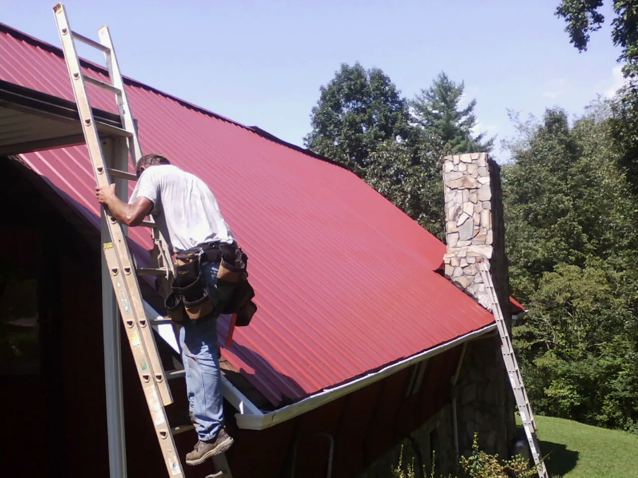 Roofer on a ladder beside a red metal roof and stone chimney — Soffit & Fascia Repair by Roofing Brothers in Kila